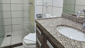 A bathroom with white tiled walls, a shower, a toilet, a sink with a granite countertop, and a mirror. - Beach Class Residence by SOLEMAR (Fortaleza)