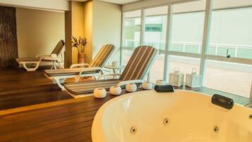 Habitación iluminada por el sol con una bañera grande, dos sillones a rayas y una pequeña mesa con objetos decorativos.