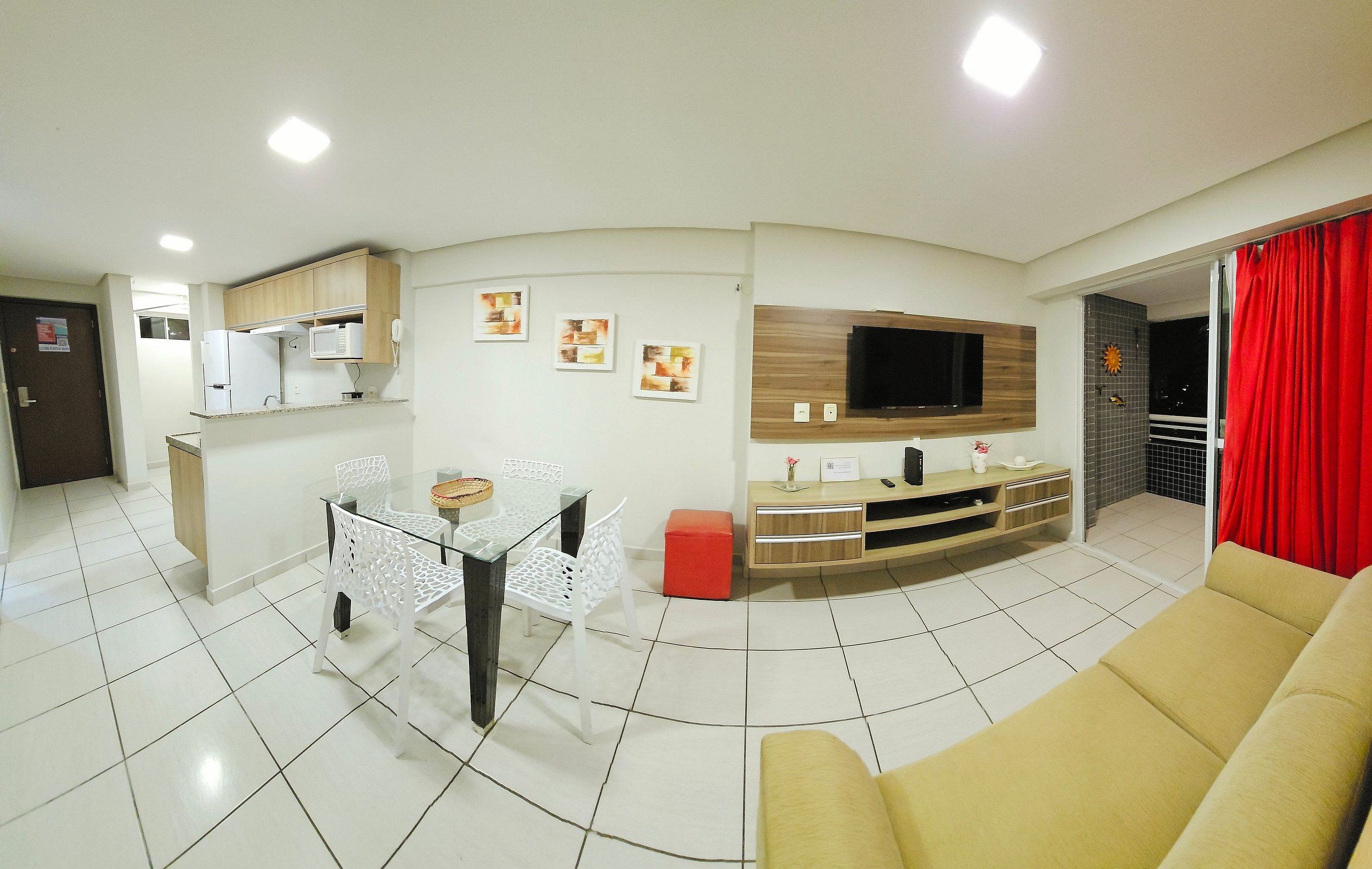 A modern living room with a sofa, a dining table with chairs, a kitchen area with cabinets, a TV mounted on the wall, and a red curtain.