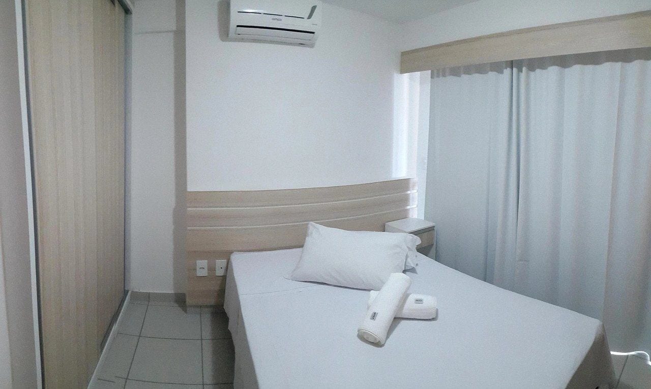 Habitación de hotel con cama, almohada, control remoto, unidad de aire acondicionado y ventana con cortinas.