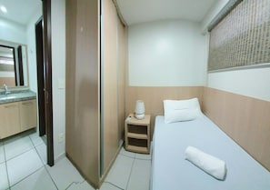 Habitación de hotel con cama, mesita de noche con lámpara, espejo, baño con lavamanos y ventana con persianas.