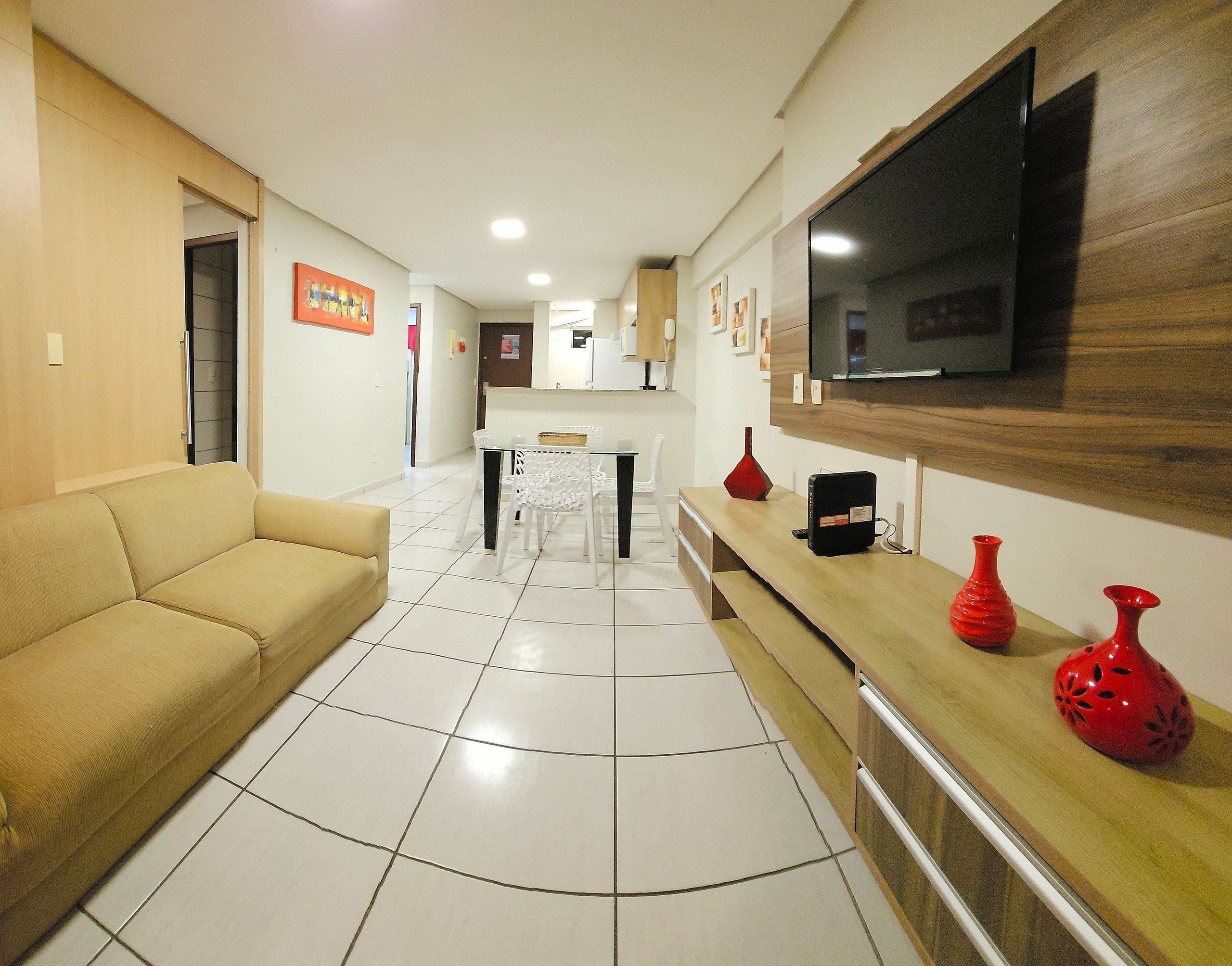 A living room with a sofa, a TV mounted on the wall, a dining area with a table and chairs, and a kitchen area in the background.