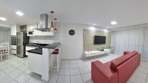 A modern kitchen with a built-in oven, microwave, and stainless steel refrigerator. There is a dining area with a white table and chairs, and a red sofa in the living room. - Beach Class Residence by SOLEMAR (Fortaleza)