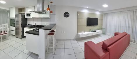 Cocina moderna con horno empotrado, microondas y refrigerador de acero inoxidable. Hay un comedor con una mesa y sillas blancas, y un sofá rojo en la sala de estar.