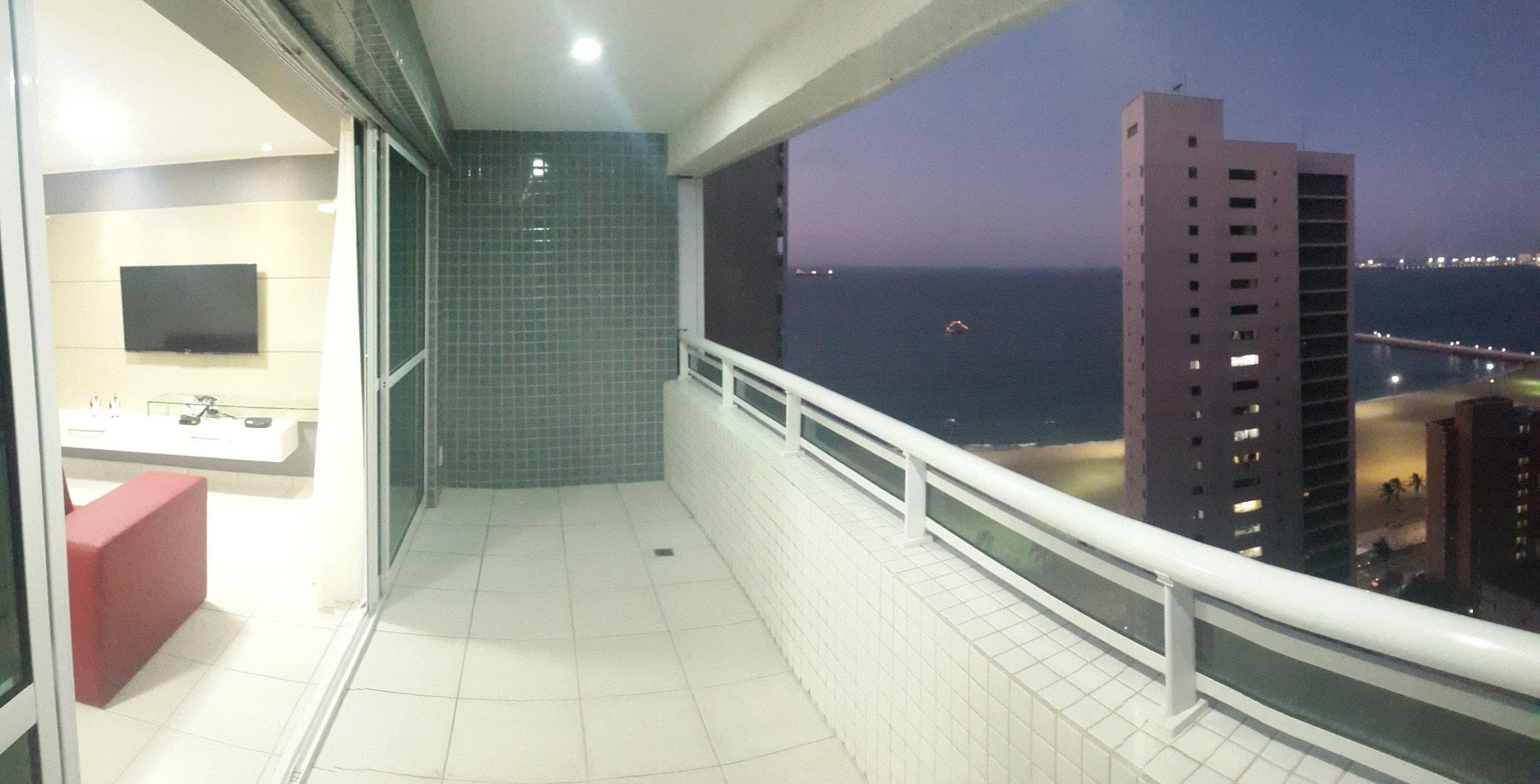 A balcony with a view of the sea and buildings, a TV mounted on the wall, and a red bench.