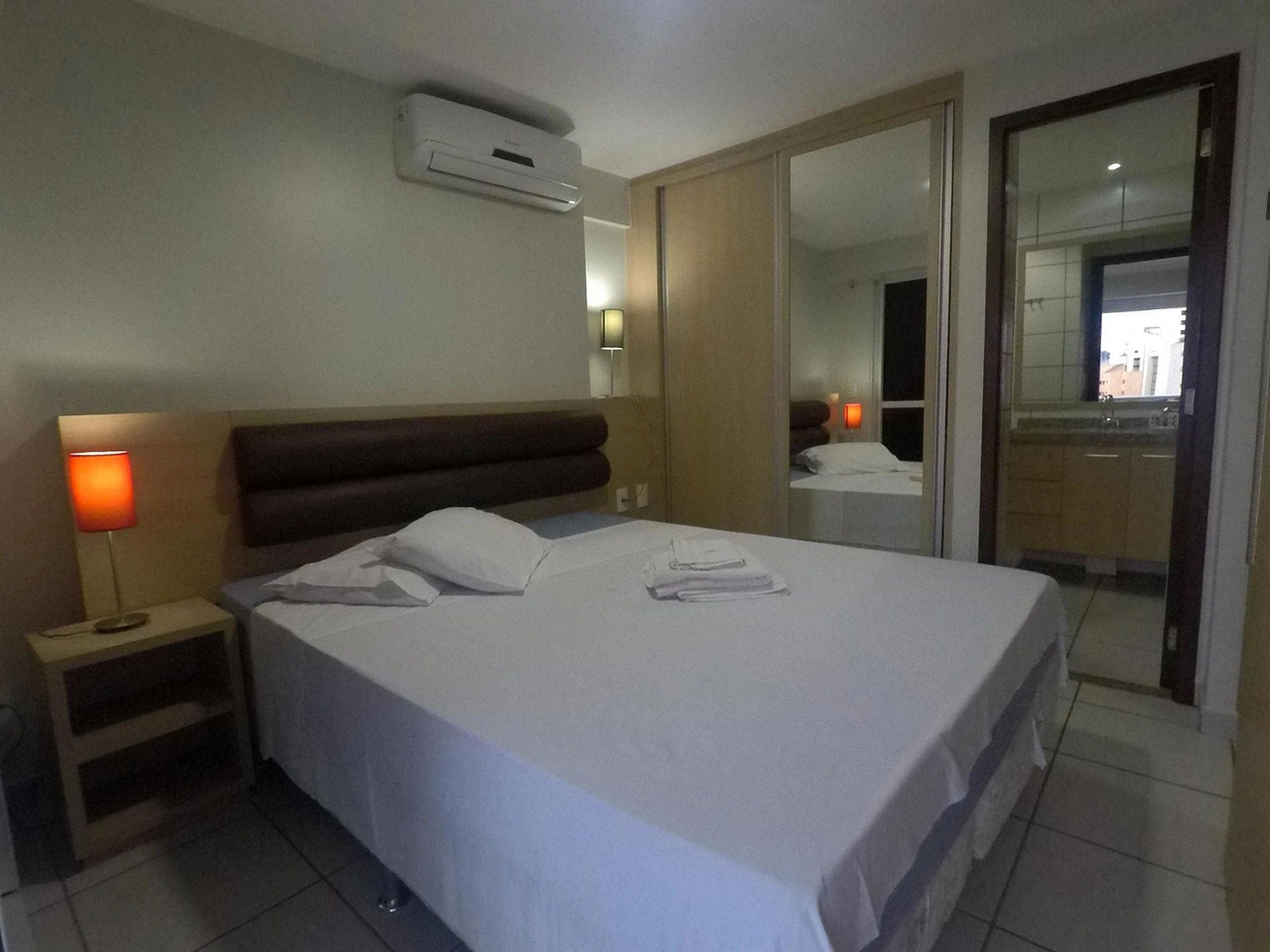 Quarto de hotel com cama, mesa de cabeceira, luminária, aparelho de ar condicionado e um espelho refletindo um banheiro.