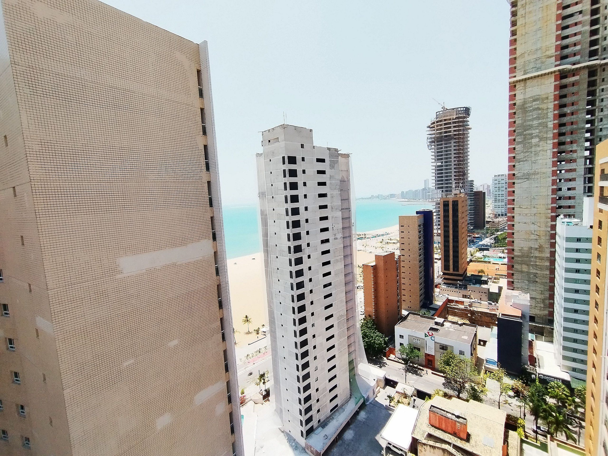 A high-rise building with a grid-like facade, a view of a beach, and other surrounding buildings.