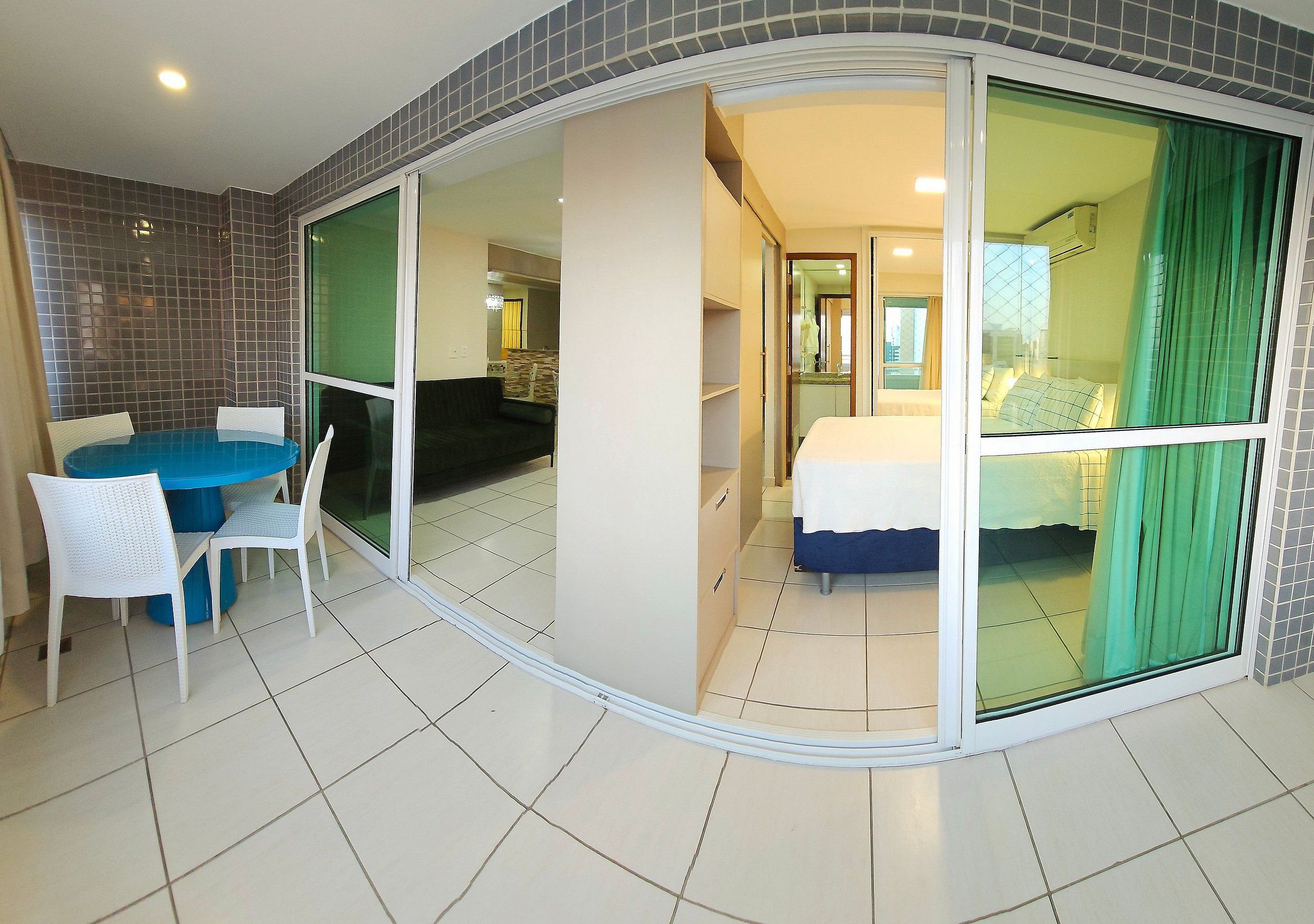 Quarto com cama, mesa e cadeiras, porta de vidro deslizante e vista para outro quarto.