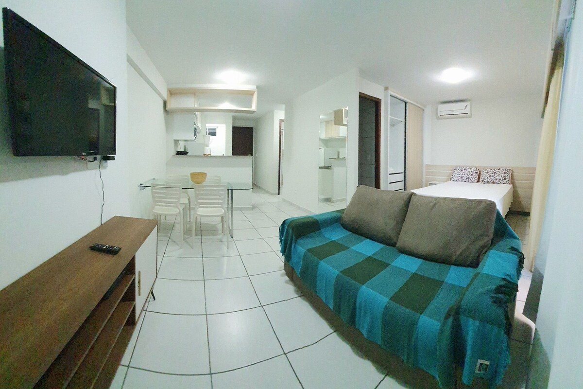 A compact hotel room with a sofa, a TV, a dining table, and a kitchenette.