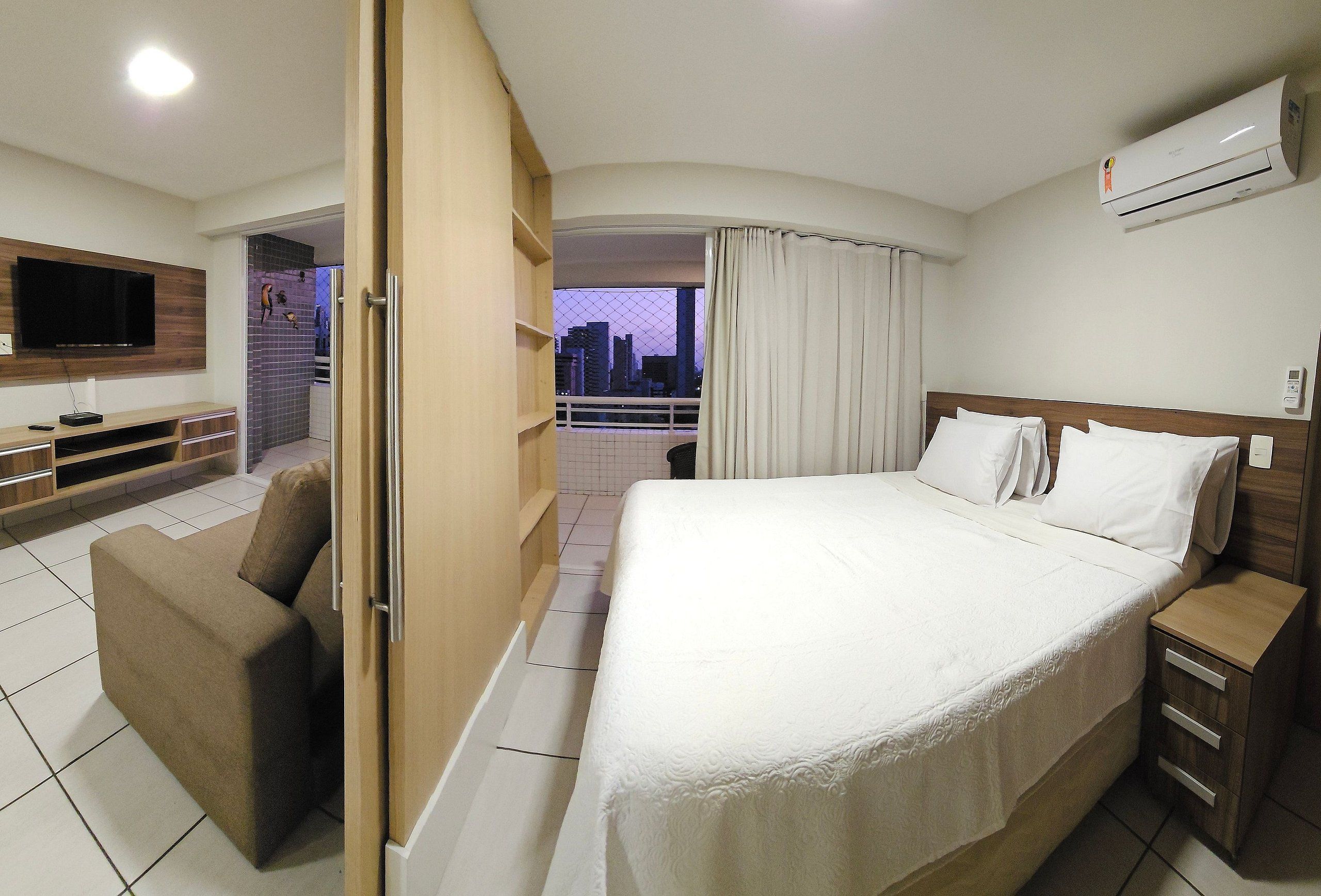 Quarto de hotel com cama, televisão, sofá e varanda com vista para a cidade.