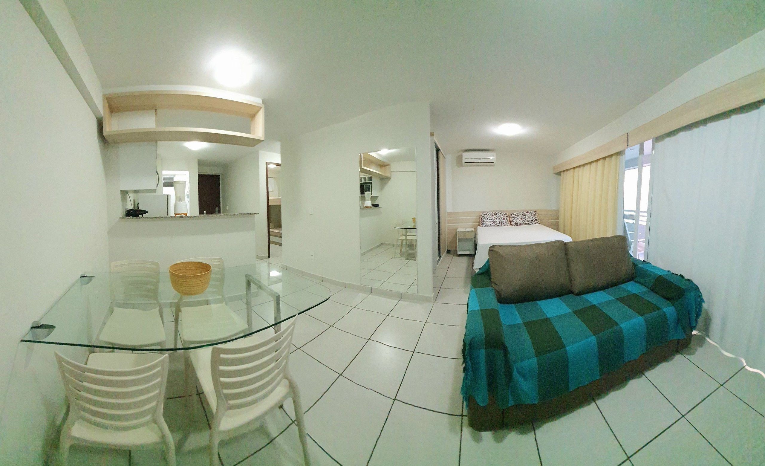 Habitació d'hotel amb taula de dinar de vidre, cadires blanques, sofà amb funda a quadres, llit amb coixins estampats i finestra amb cortines.