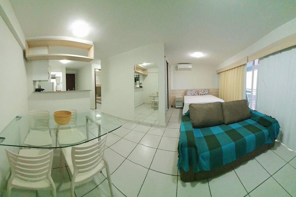 Ein Hotelzimmer mit einem Glastisch, weißen Stühlen, einem Sofa mit kariertem Bezug, einem Bett mit gemusterten Kissen und einem Fenster mit Vorhängen.