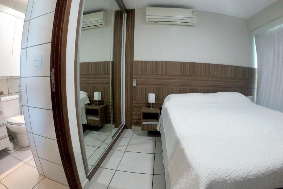 A hotel room with a bed, a bedside table, a mirror, a toilet, and an air conditioning unit.