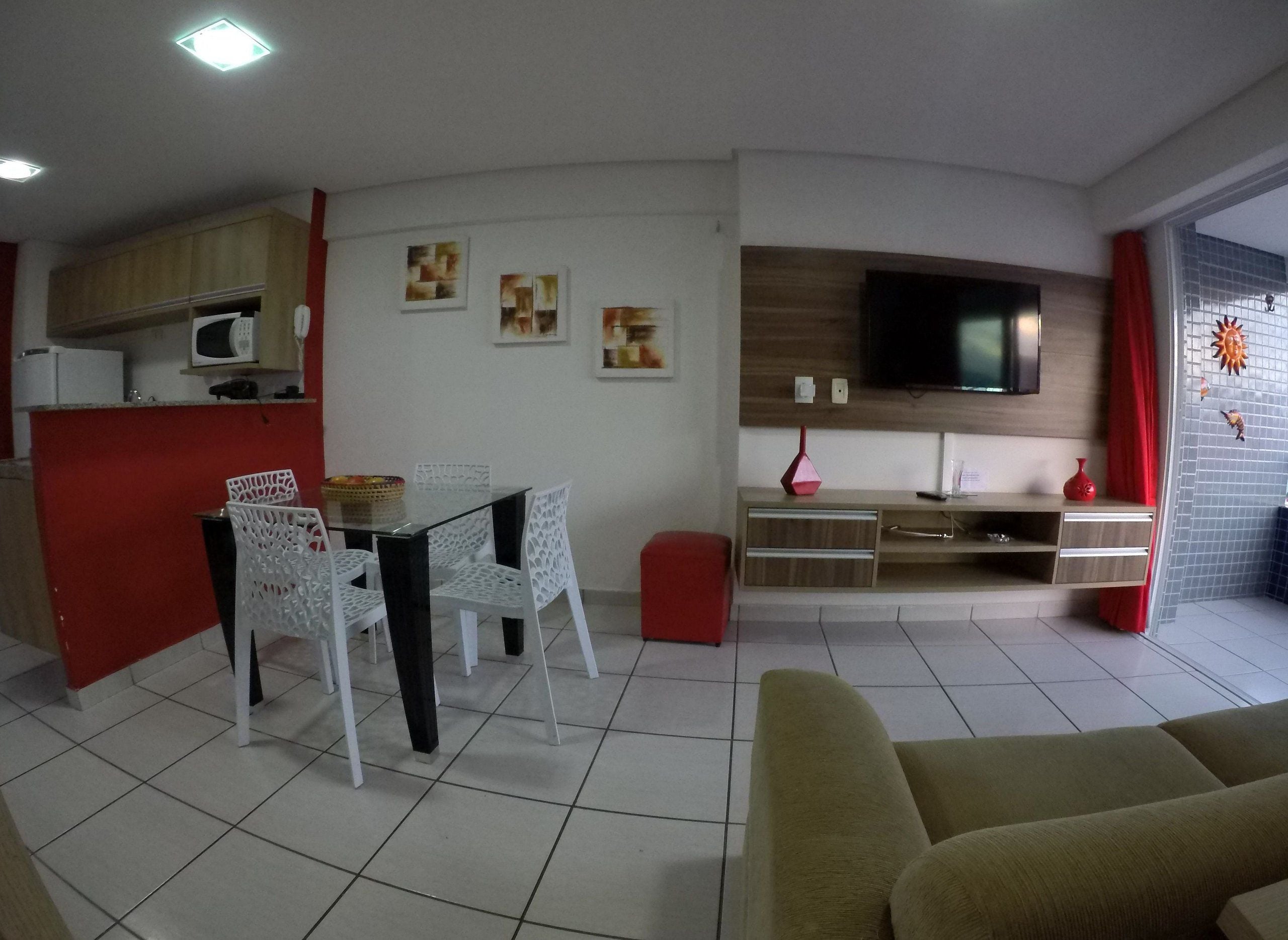 A living room with a sofa, a dining table with chairs, a TV mounted on the wall, and a kitchen area with cabinets and a microwave.