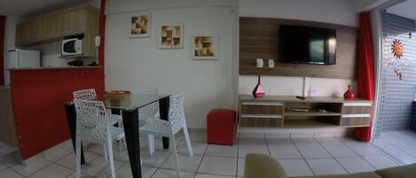 A living room with a sofa, a dining table with chairs, a TV mounted on the wall, and a kitchen area with cabinets and a microwave.