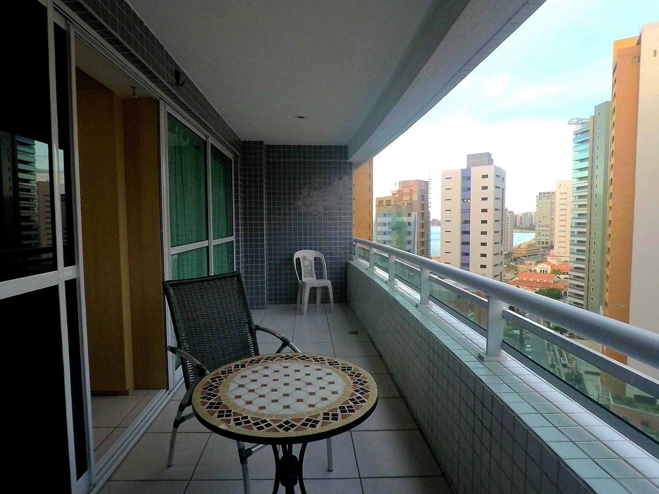 Un balcón con una mesa y sillas de cerámica, vista a edificios de la ciudad y barandilla de vidrio.