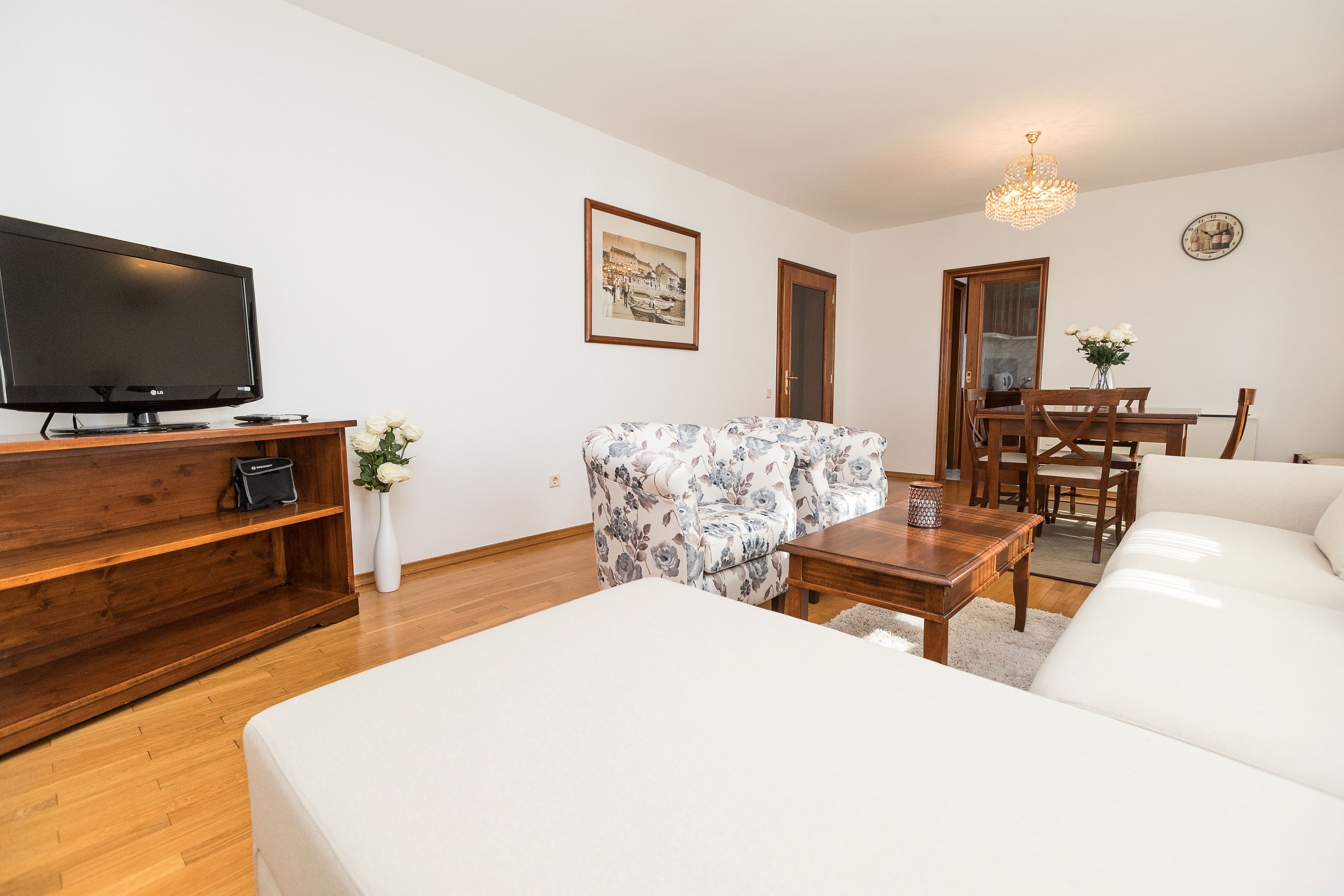 A modern living room with a flat-screen TV, a floral sofa, a wooden dining table, and a white couch.