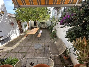 Eine überdachte Terrasse mit Holztisch und Bänken, umgeben von Kübelpflanzen und einem kleinen Garten.