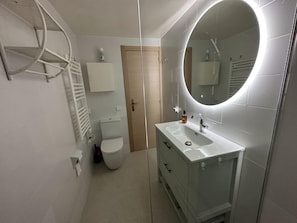 Ein modernes Badezimmer mit einem weißen Waschbecken, einem Spiegel mit runder Beleuchtung, einer Toilette und einem Handtuchhalter.