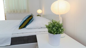 A neatly made bed with a pillow, a bedside plant, and a lamp on a white nightstand. - Apartments Casa Mia (Crikvenica)