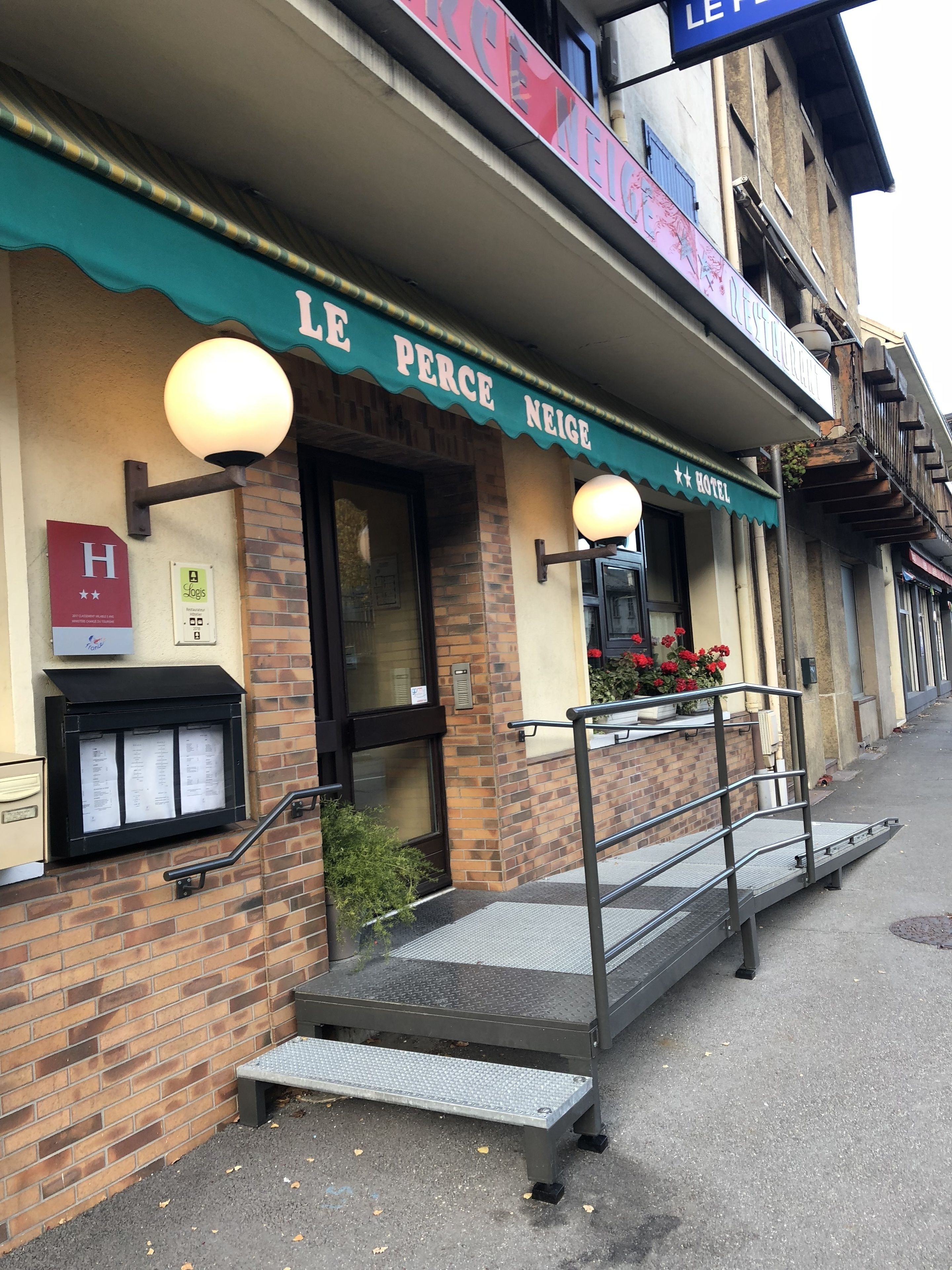 A hotel named "Le Perce Neige" with a brick facade, a green awning, and a small outdoor seating area with a metal railing.