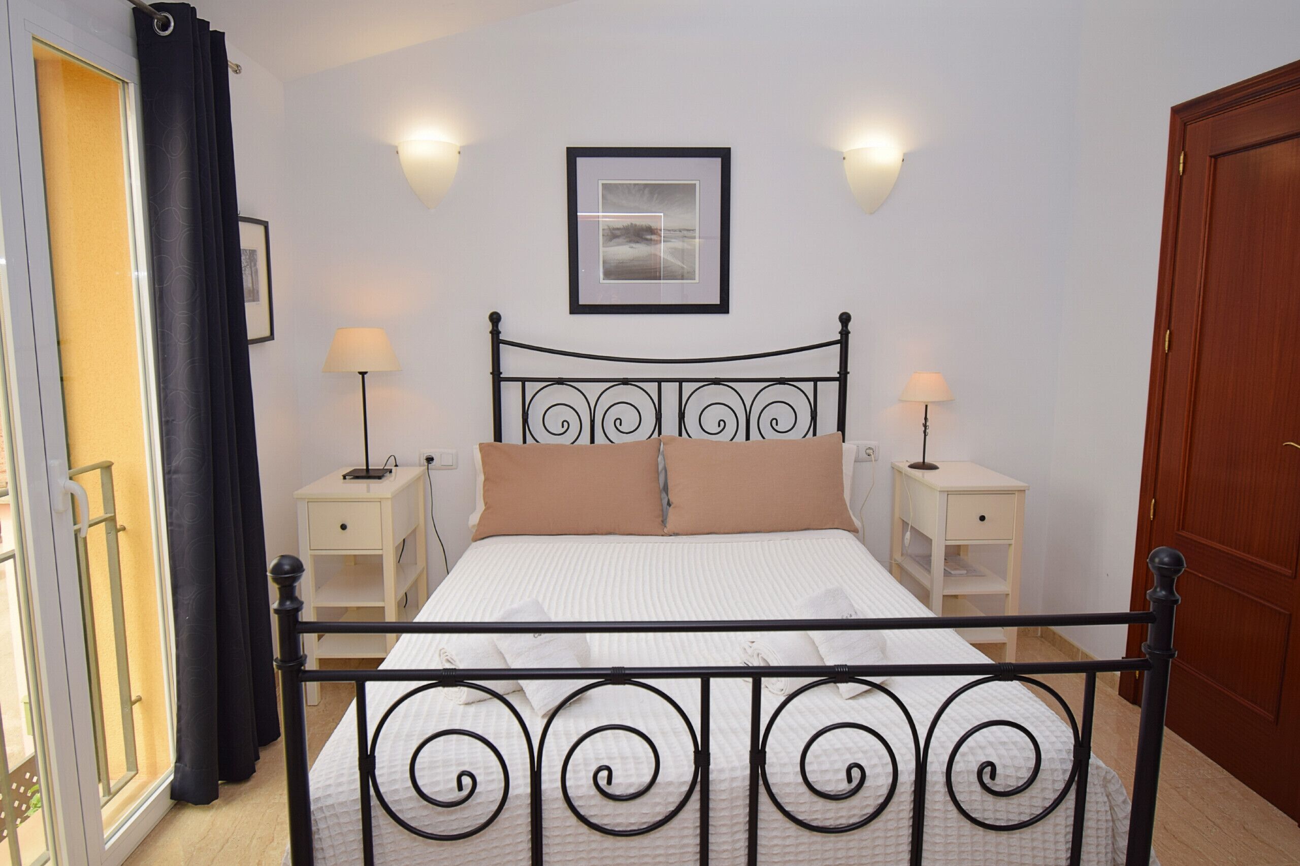 Une chambre à coucher avec un lit, deux tables de chevet avec des lampes, un tableau encadré au mur et une porte.