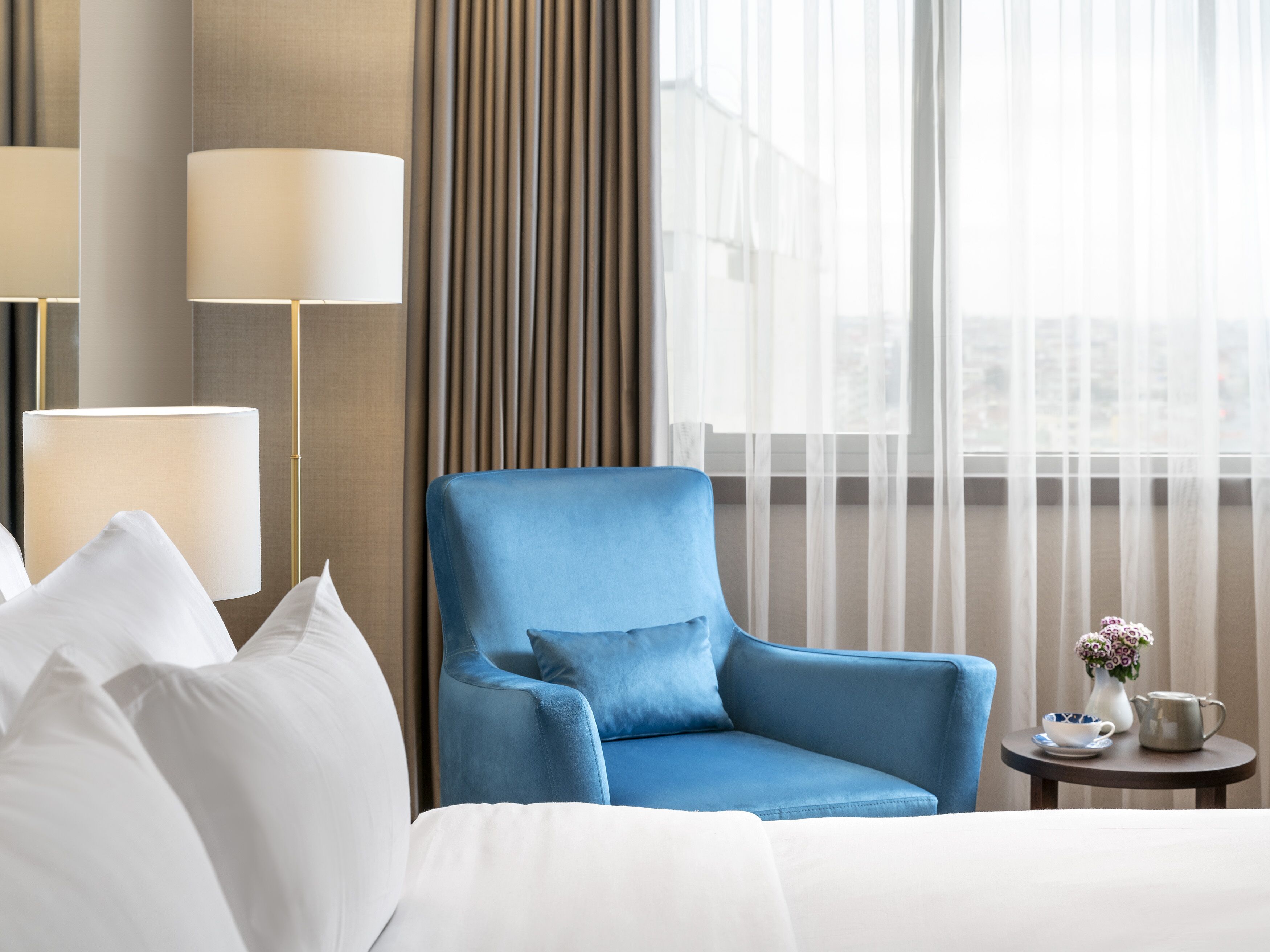 Una camera d'albergo con una poltrona blu, un letto con lenzuola bianche, un tavolino rotondo con un vaso e un teiera, e un'ampia finestra con tende trasparenti.