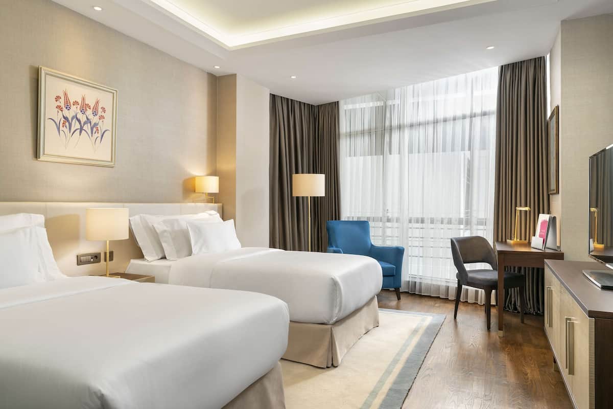 A hotel room with two beds, a desk, a chair, a TV, and a large window with curtains.