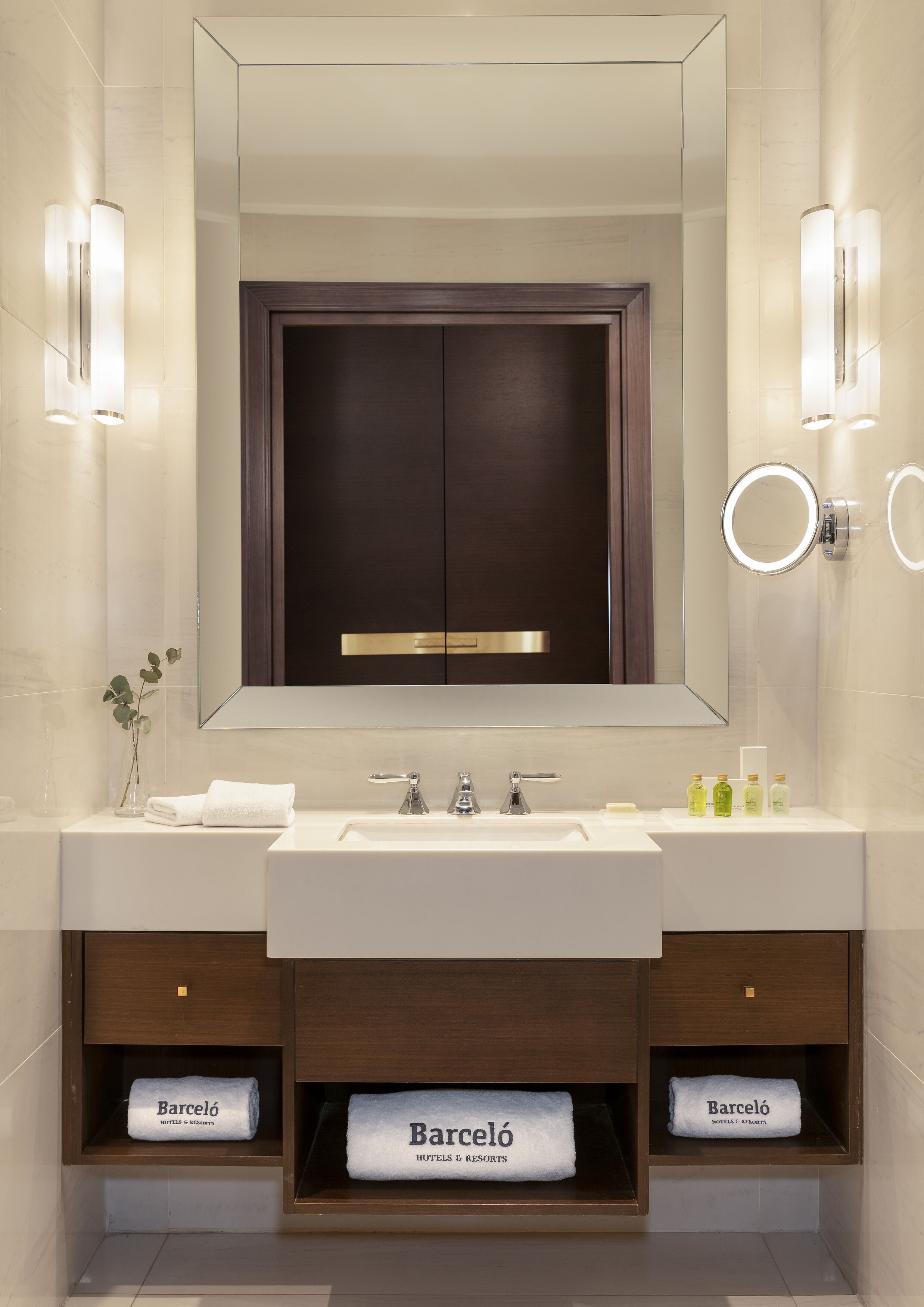 A modern bathroom with a large mirror, two sinks, and a vanity with shelves holding towels.