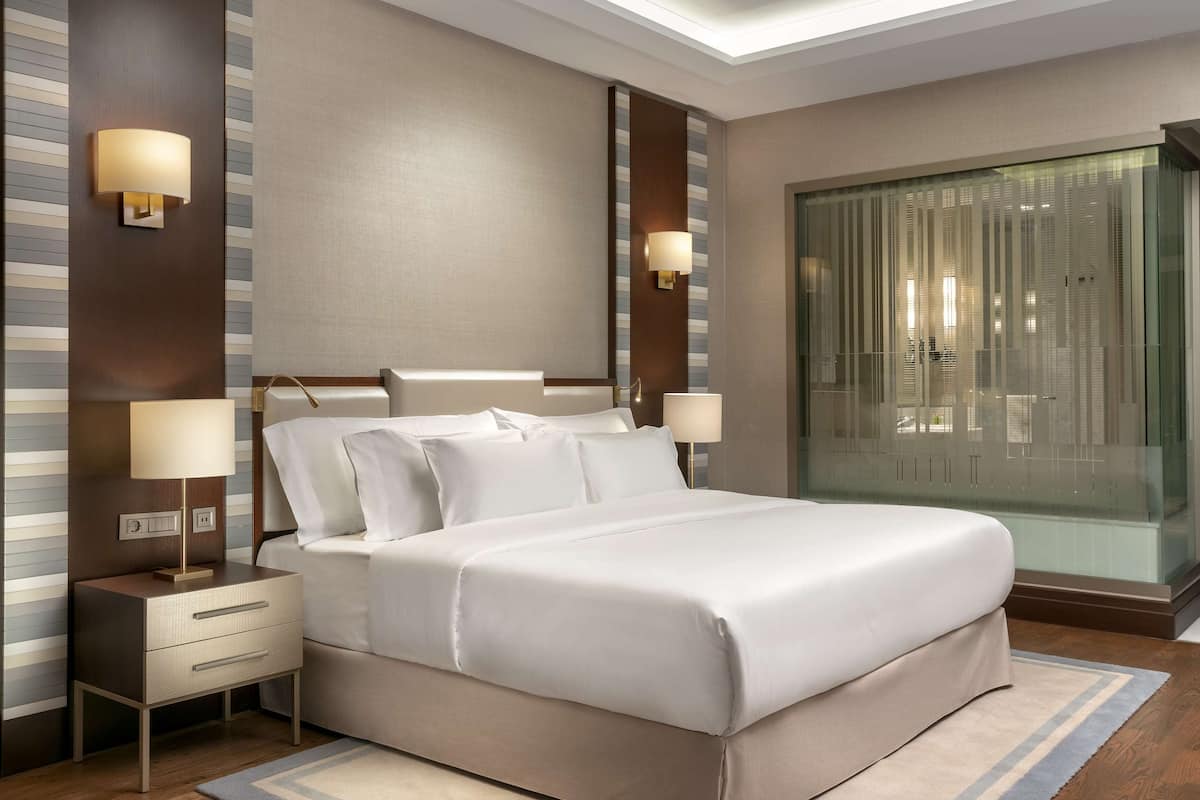 A modern hotel room with a large bed, bedside lamps, a nightstand, and a glass partition wall.