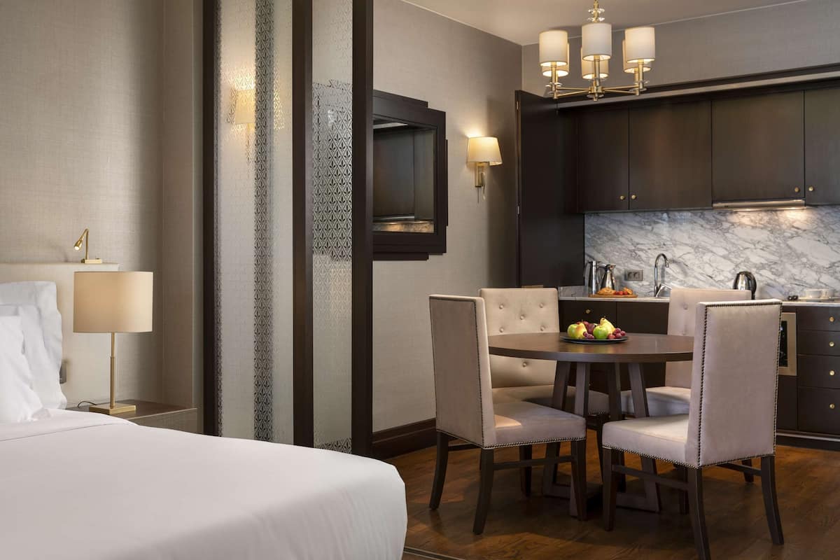 A hotel room with a bed, a desk, a dining table with chairs, a kitchen area with a sink and cabinets, and a TV mounted on the wall.