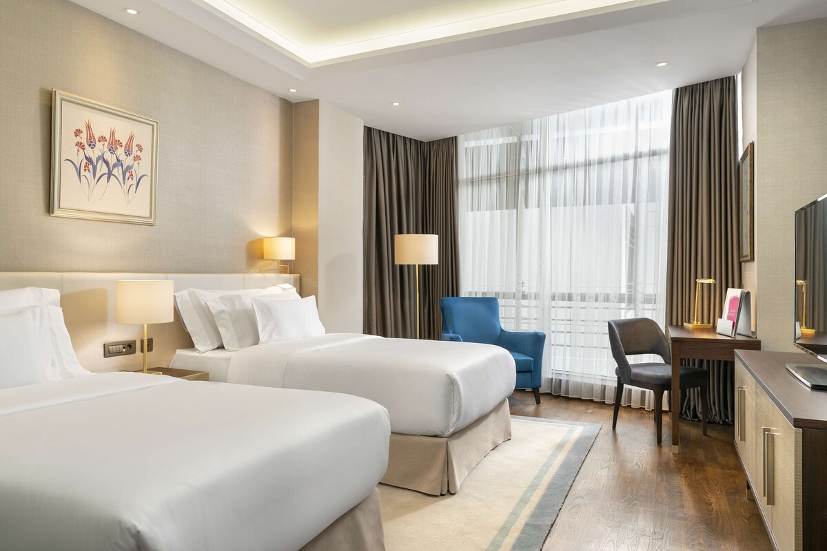 A hotel room with two beds, a desk, a chair, a TV, and a large window with curtains.