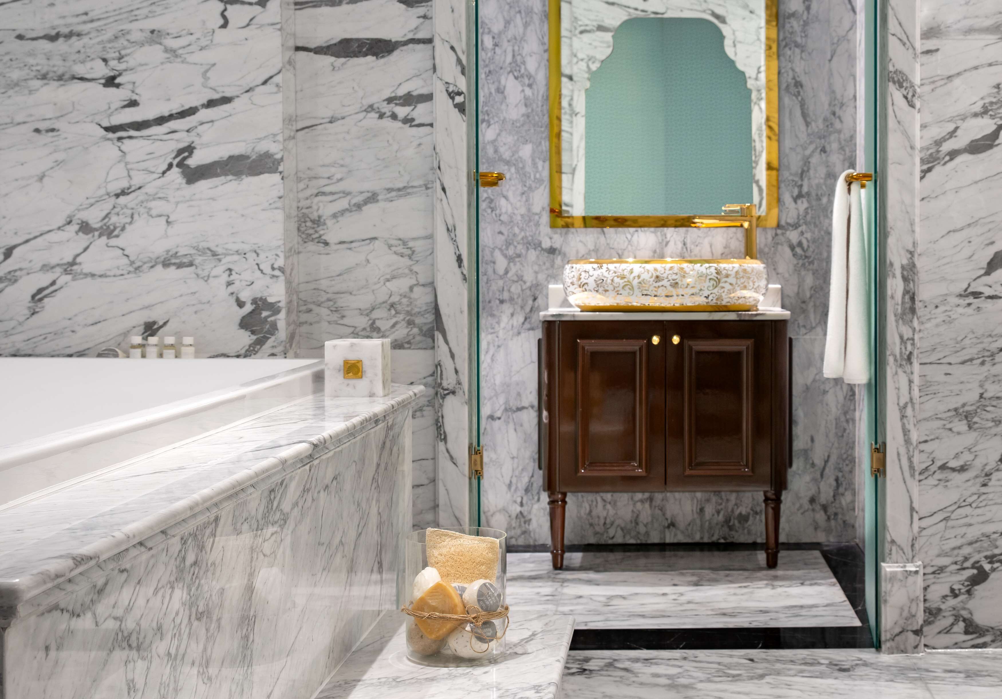 A marble bathroom with a bathtub, a vanity with a mirror, and a glass jar with toiletries.