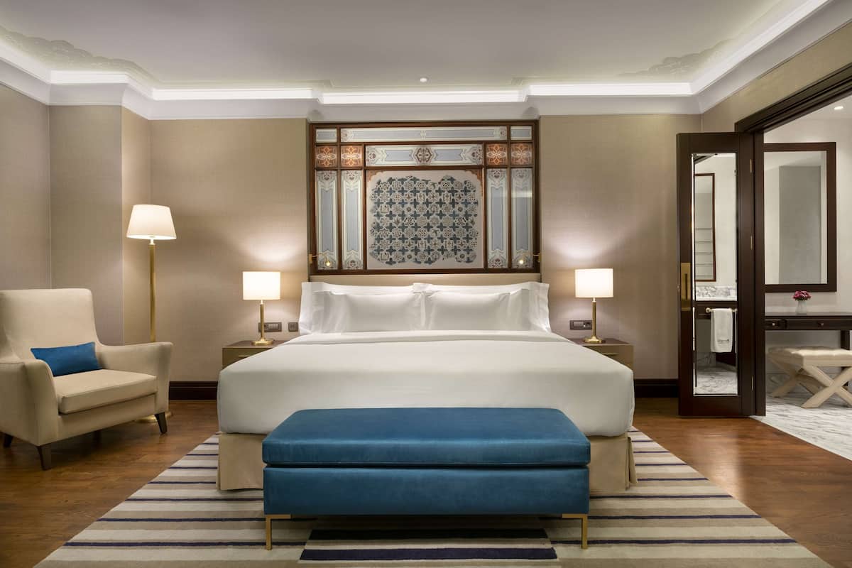 A hotel room with a large bed, two bedside tables with lamps, a chair, a blue bench, a decorative wall panel, and a mirror.