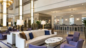 Un vestíbulo amplio con techo alto, múltiples candelabros, un área de estar central con sofás y sillones, y una recepción al fondo.