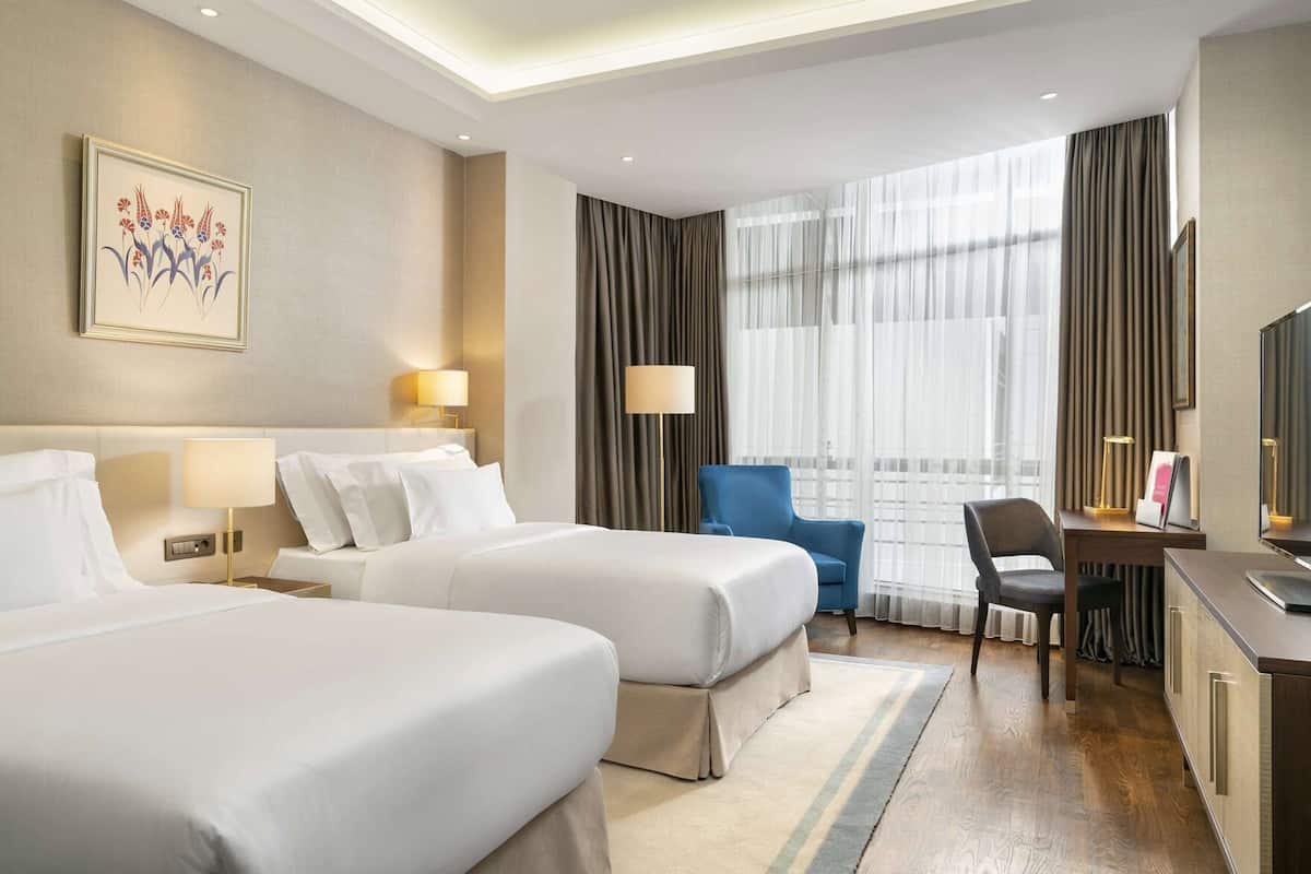 A hotel room with two beds, a desk, a chair, a TV, and a large window with curtains.