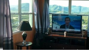 Un televisor mostra un informatiu, una làmpada i una vista de muntanyes per la finestra.