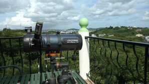 A telescope labeled 'CELESTRON AstroMaster 114' mounted on a platform with a railing, overlooking a green landscape with houses in the distance. - Cook's Estate (St. John's)