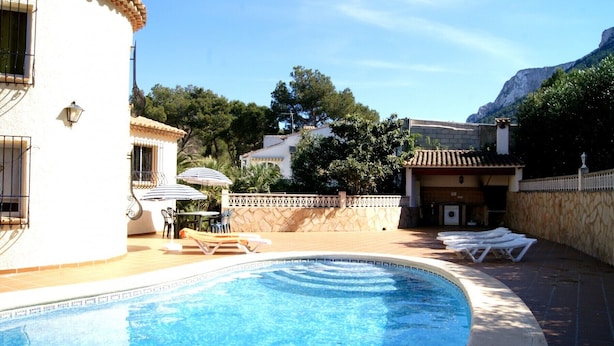 A swimming pool with lounge chairs and umbrellas, surrounded by a stone wall and vegetation. - 0538 Villa Efaistos en el Montgo (Denia)
