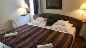 Una cama bien hecha con una colcha a rayas, dos lámparas de noche, un radiador y una obra enmarcada en la pared.