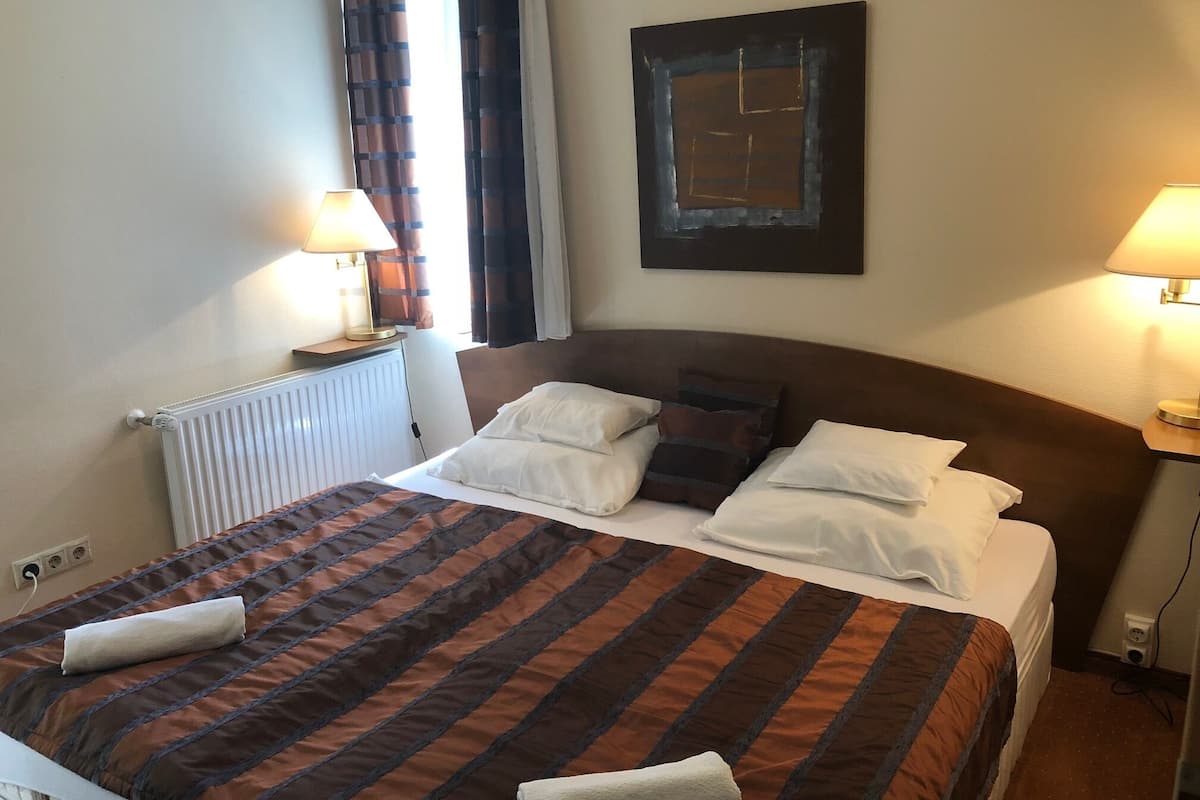 Un lit bien fait, recouvert d’une couvre-lit à rayures, deux lampes de chevet, un radiateur et une œuvre d’art encadrée accrochée au mur.