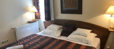 A neatly made bed with a striped bedspread, two bedside lamps, a radiator, and a framed artwork on the wall.