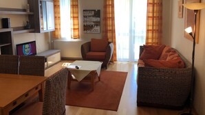 Una sala de estar con muebles de mimbre, una mesa y un sofá. Hay un televisor sobre un mueble bajo, una estantería y una ventana con cortinas.