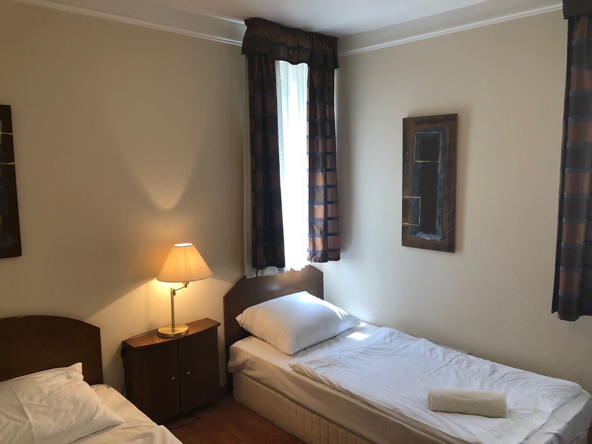 Een hotelkamer met een enkel bed, een nachtkastje met een lamp, een raam met gordijnen en een lijst met een afbeelding aan de muur.