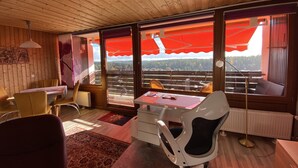 Et værelse med trævægge og -loft, et spiseområde med gule stole, et hvidt skrivebord med stol, samt store vinduer med røde markiser.