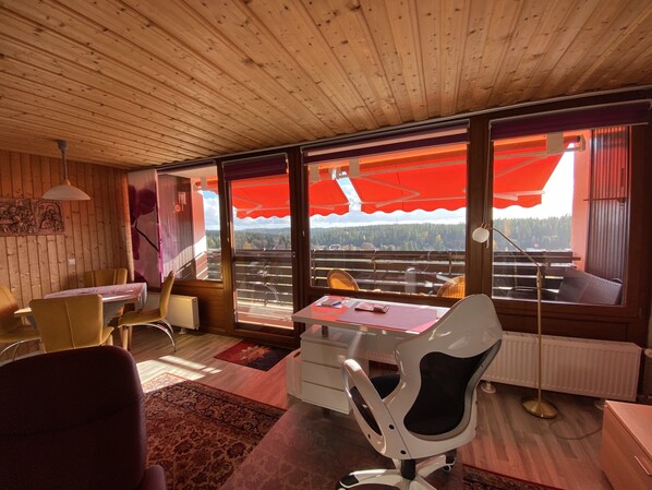 A room with wooden walls and ceiling, a dining area with yellow chairs, a white desk with a chair, and large windows with red awnings. - North Black Forest 2-room apartment 58 sqm, Dieter Hoffmann, Freudenstadt-Kniebis (Kniebis)