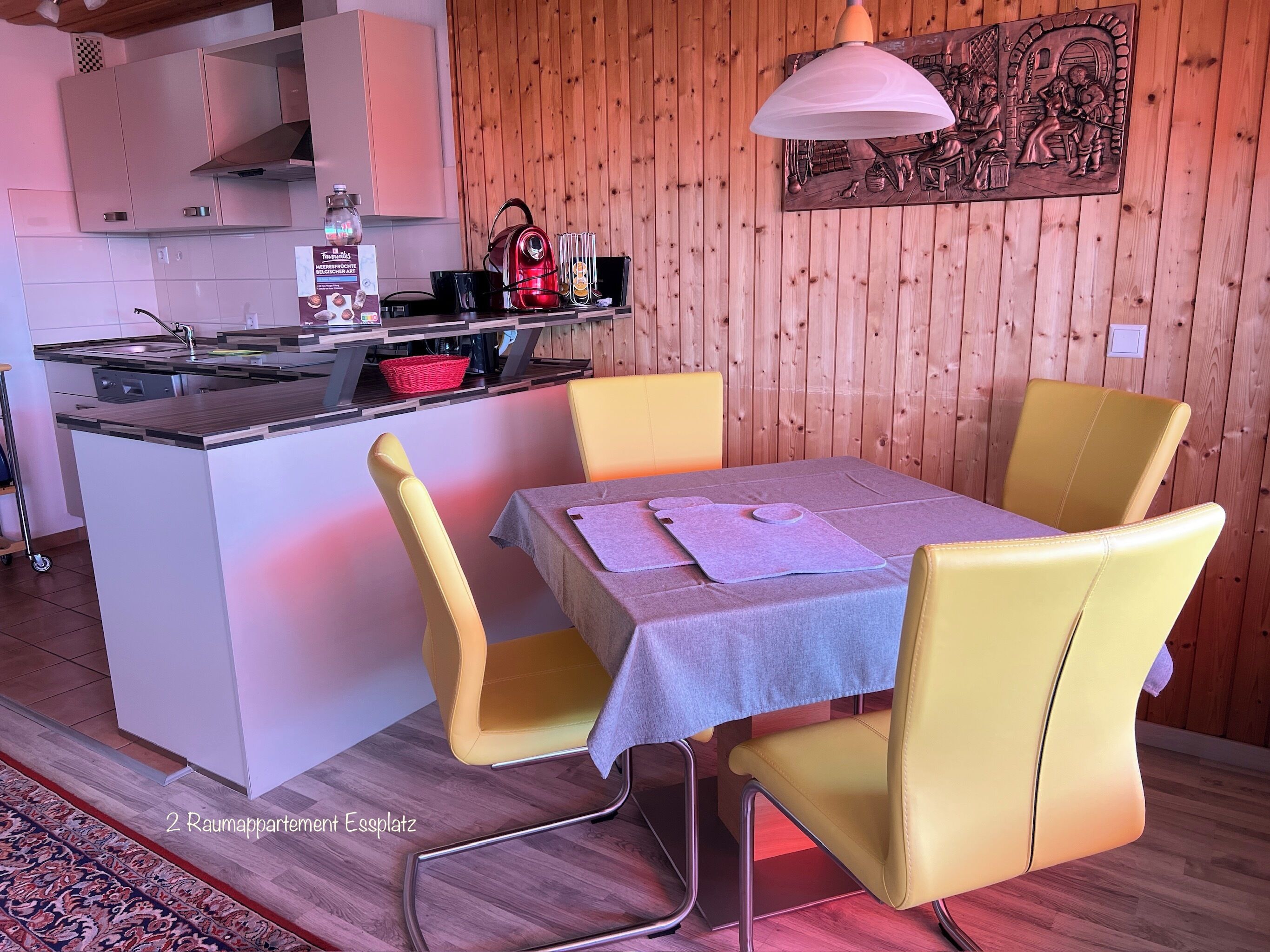 A compact kitchen with a central island, a dining table with yellow chairs, and a wooden wall with a mural.