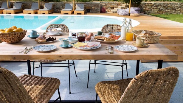 A poolside breakfast setup with wicker chairs, a wooden table, and a pool in the background.