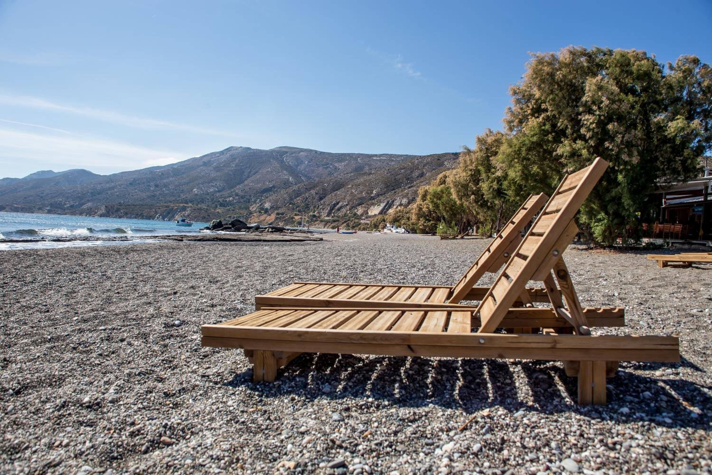 Una silla reclinable de madera en una playa de piedras con vistas al mar y a las montañas.