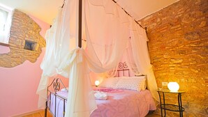 A bedroom with a four-poster bed, a stone wall, and a small table with a lamp.